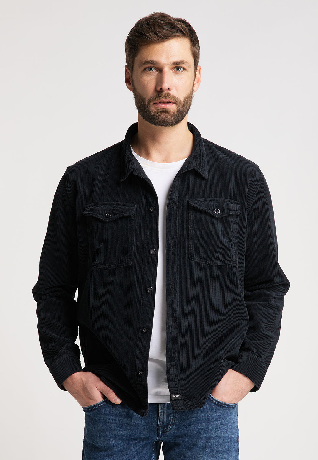 Ein Mann mit kurzen braunen Haaren und Bart trägt eine schwarze Hemdjacke Cord Originals von Recaro_Merch_Daniel über einem weißen T-Shirt und blauen Jeans und steht mit den Händen in den Taschen vor einem schlichten hellen Hintergrund.