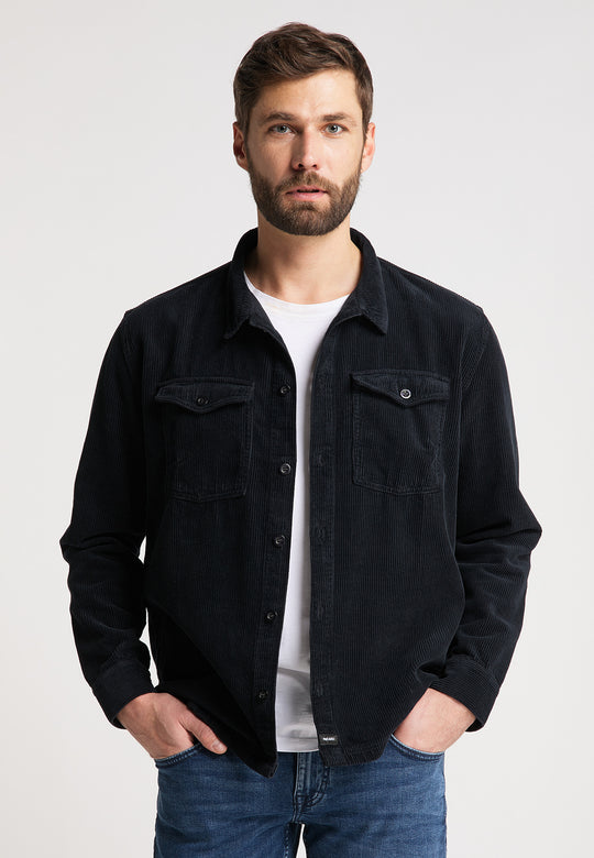 Ein Mann mit kurzen braunen Haaren und Bart trägt eine schwarze Hemdjacke Cord Originals von Recaro_Merch_Daniel über einem weißen T-Shirt und blauen Jeans und steht mit den Händen in den Taschen vor einem schlichten hellen Hintergrund.