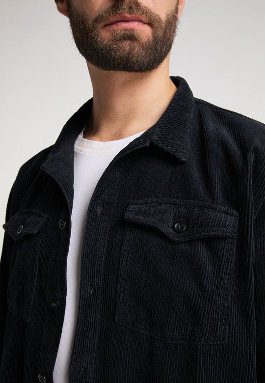 Eine Nahaufnahme eines Mannes, der eine schwarze Recaro_Merch_Daniel Hemdjacke Cord Originals über einem weißen T-Shirt trägt, wodurch sein Bart, seine Lippen und sein Oberkörper vor einem schlichten Hintergrund hervorgehoben werden.
