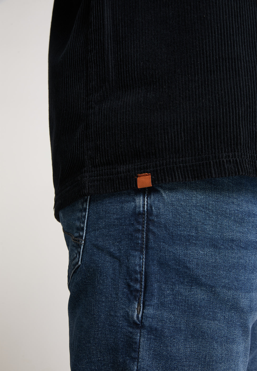 Person in Blue-Denim-Jeans und schwarzem Rippenhemd mit kleinem braunen Etikett, gestylt mit der Recaro Hemdjacke Cord Originals, fotografiert von der Taille abwärts vor einem schlichten Hintergrund.