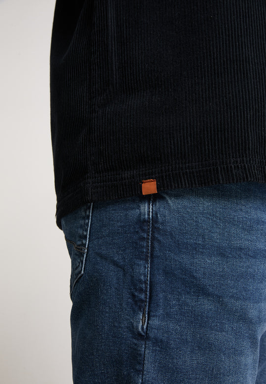 Person in Blue-Denim-Jeans und schwarzem Rippenhemd mit kleinem braunen Etikett, gestylt mit der Recaro Hemdjacke Cord Originals, fotografiert von der Taille abwärts vor einem schlichten Hintergrund.