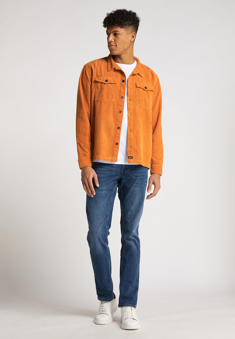 Ein junger Mann steht leicht gedreht da und trägt eine leuchtend orange Hemdjacke Cord Originals von RECARO über einem weißen T-Shirt, Bluejeans und weißen Turnschuhen vor einem schlichten hellen Hintergrund.