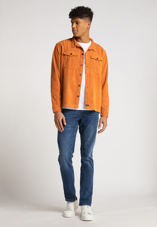 Ein junger Mann steht leicht gedreht da und trägt eine leuchtend orange Hemdjacke Cord Originals von RECARO über einem weißen T-Shirt, Bluejeans und weißen Turnschuhen vor einem schlichten hellen Hintergrund.