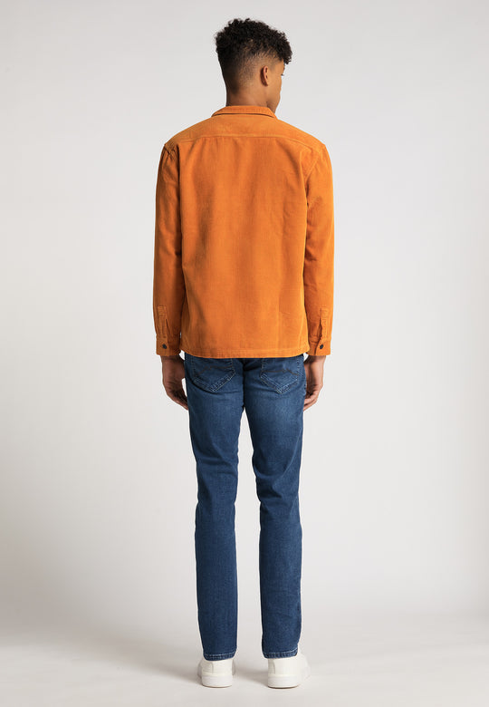 Eine Person steht mit dem Rücken zur Kamera und trägt eine orangefarbene RECARO Hemdjacke Cord Originals, Bluejeans und weiße Turnschuhe vor einem schlichten hellen Hintergrund.