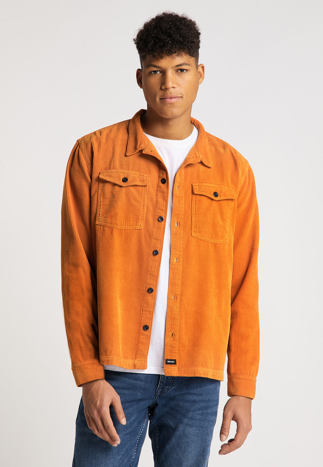 Ein junger Mann mit kurzen lockigen Haaren trägt die RECARO Hemdjacke Cord Originals in Orange über einem weißen T-Shirt und blauen Jeans und steht mit dem Gesicht nach vorne vor einem schlichten hellen Hintergrund.