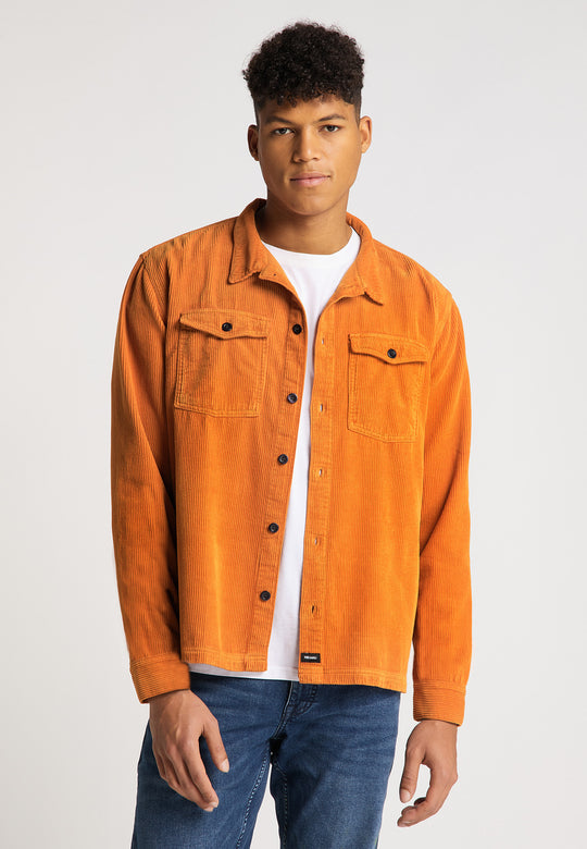 Ein junger Mann mit kurzen lockigen Haaren trägt die RECARO Hemdjacke Cord Originals in Orange über einem weißen T-Shirt und blauen Jeans und steht mit dem Gesicht nach vorne vor einem schlichten hellen Hintergrund.
