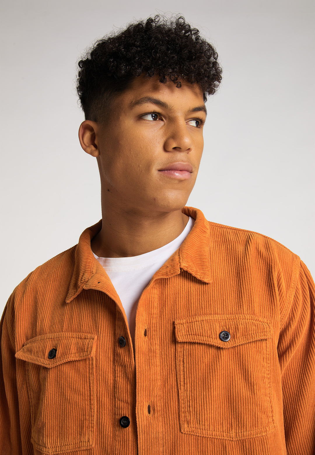 Ein junger Mann mit kurzen, lockigen Haaren trägt eine Recaro_Merch_Daniel Hemdjacke Cord Originals in Orange über einem weißen Hemd und blickt vor einem schlichten hellen Hintergrund zur Seite.
