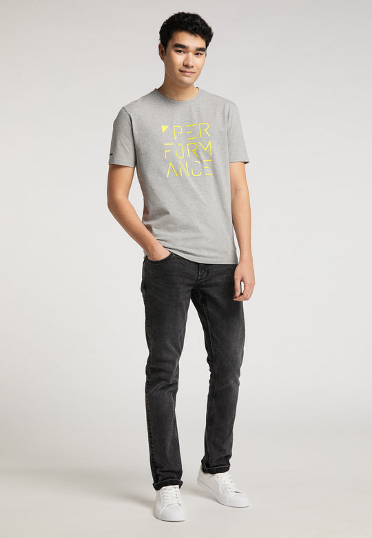 Ein junger Mann mit kurzen dunklen Haaren steht vor einem schlichten Hintergrund. Er trägt das graue Recaro_Merch_Daniel T-Shirt Performance mit gelbem „PERFORMANCE“-Schriftzug, schwarze Jeans und weiße Turnschuhe. Er sieht entspannt aus und hat eine Hand in der Tasche.