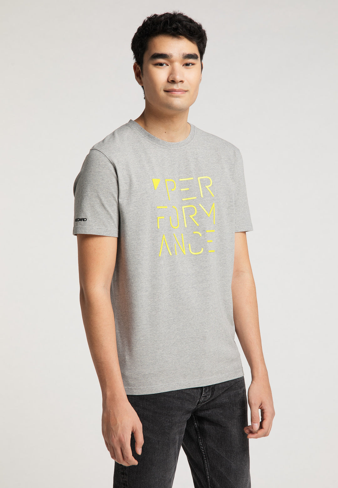 Ein junger Mann mit kurzen dunklen Haaren trägt schwarze Jeans und ein graues Recaro_Merch_Daniel T-Shirt Performance mit PERFORMANCE-PRINT in Gelb auf der Vorderseite und steht vor einem schlichten hellen Hintergrund.