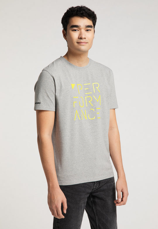 Ein junger Mann mit kurzen dunklen Haaren trägt schwarze Jeans und ein graues Recaro_Merch_Daniel T-Shirt Performance mit PERFORMANCE-PRINT in Gelb auf der Vorderseite und steht vor einem schlichten hellen Hintergrund.