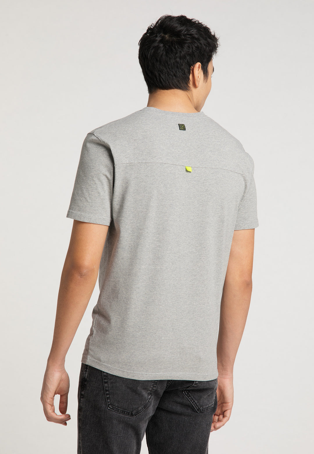 Eine Person blickt von der Kamera weg und trägt das hellgraue Recaro_Merch_Daniel T-Shirt Performance mit Performance-Print-Logo und dunkle Jeans. Der lockere Schnitt ist vor einem schlichten, hellen Hintergrund zu sehen.