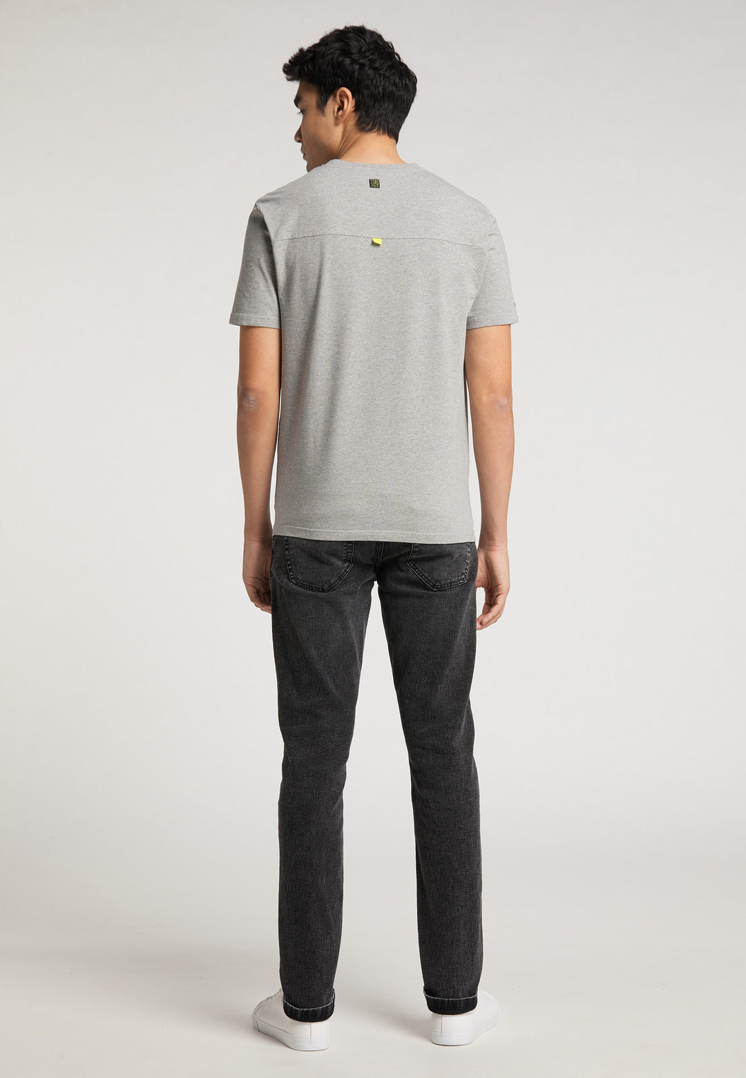 Ein Mann blickt von der Kamera weg und trägt ein hellgraues Recaro_Merch_Daniel T-Shirt Performance mit einem dezenten RECARO-Logo, dazu dunkle Jeans und weiße Sneakers vor einem schlichten hellen Hintergrund.