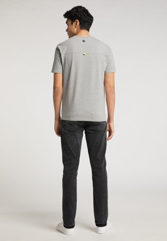 Ein Mann blickt von der Kamera weg und trägt ein hellgraues Recaro_Merch_Daniel T-Shirt Performance mit einem dezenten RECARO-Logo, dazu dunkle Jeans und weiße Sneakers vor einem schlichten hellen Hintergrund.