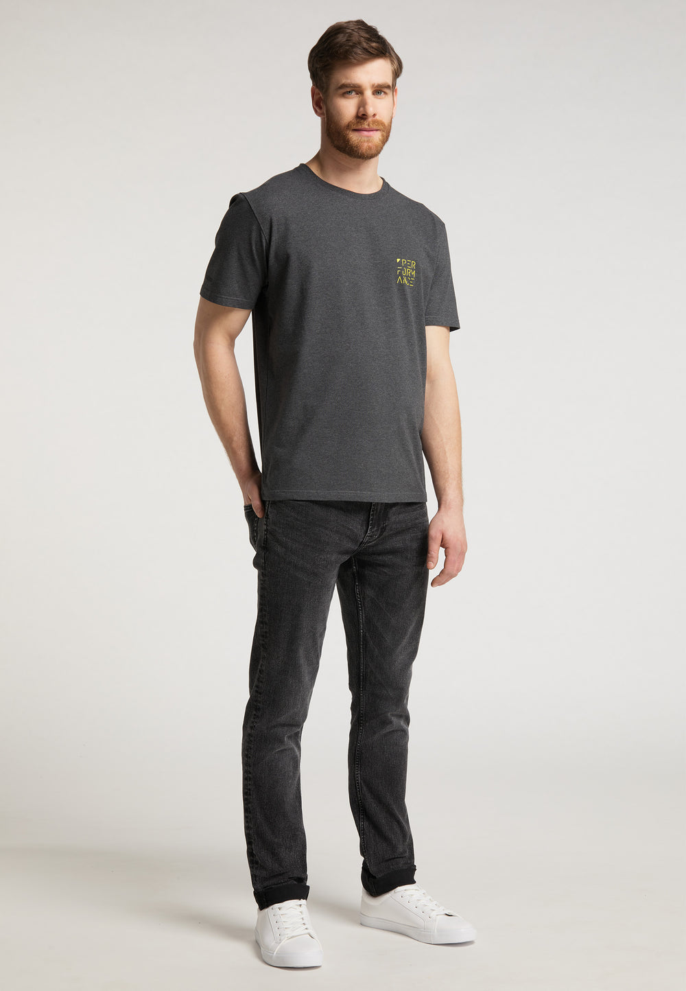 Ein Mann mit kurzen braunen Haaren und Bart posiert in einem Studio. Er trägt das Recaro_Merch_Daniel T-Shirt Performance in Dunkelgrau, schwarze Jeans und weiße Sneakers. Sein Blick ist leicht zur Seite gerichtet und sein Gesichtsausdruck neutral.