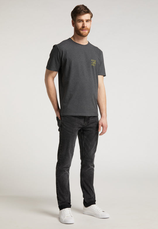 Ein Mann mit kurzen braunen Haaren und Bart posiert in einem Studio. Er trägt das Recaro_Merch_Daniel T-Shirt Performance in Dunkelgrau, schwarze Jeans und weiße Sneakers. Sein Blick ist leicht zur Seite gerichtet und sein Gesichtsausdruck neutral.