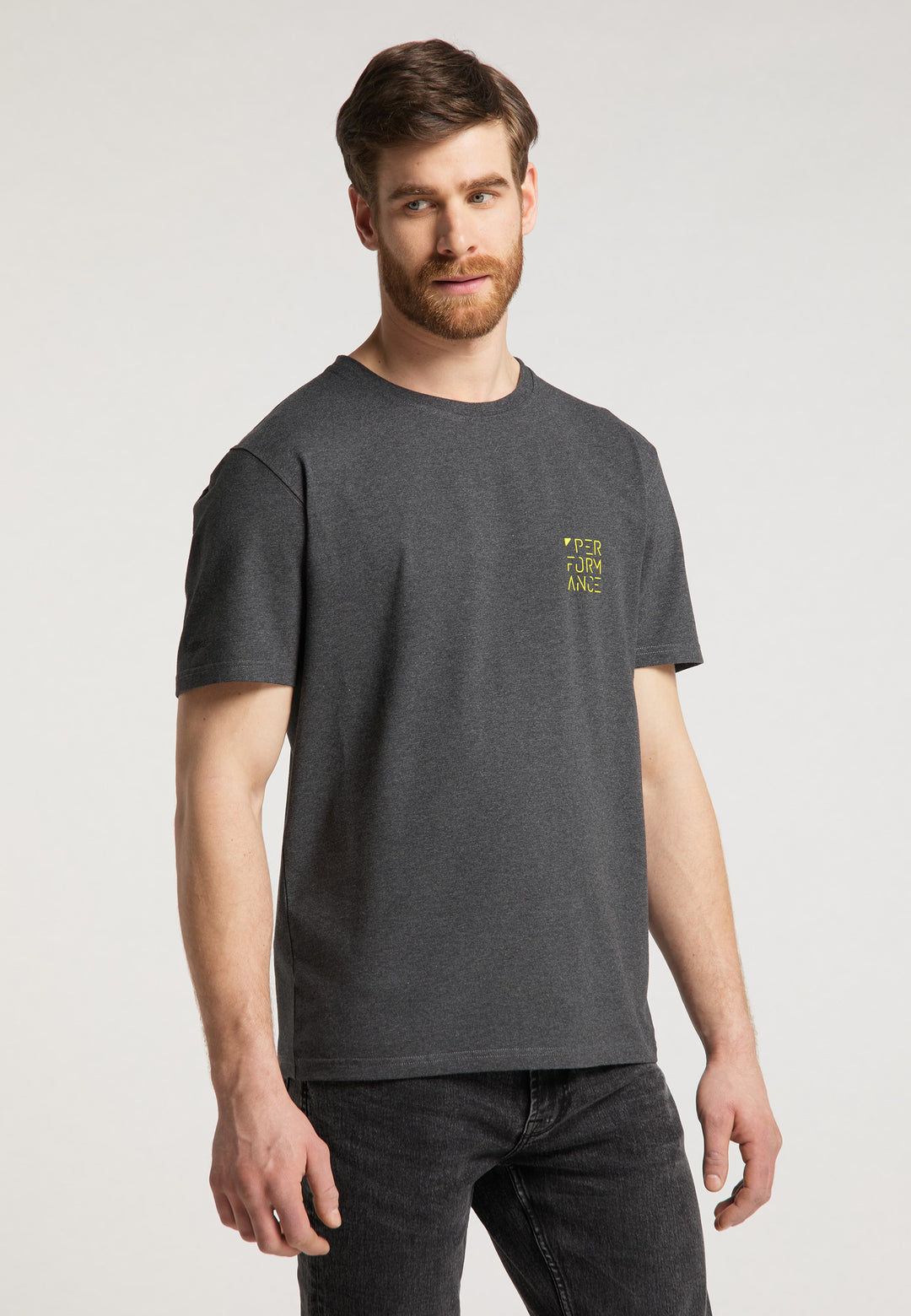 Ein bärtiger Mann mit kurzen braunen Haaren steht leicht nach links gedreht und trägt ein dunkelgraues Recaro_Merch_Daniel T-Shirt Performance und schwarze Relaxed-Fit-Jeans. Der Hintergrund ist schlicht und hell gehalten.