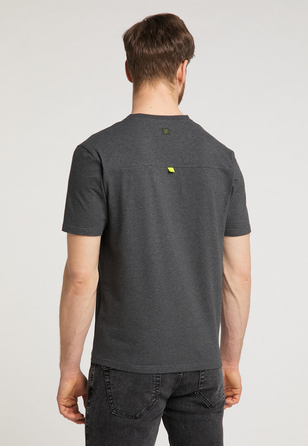 Ein Mann mit kurzen braunen Haaren, der das Recaro_Merch_Daniel T-Shirt Performance in Relaxed-Fit Dunkelgrau mit dezentem RECARO Logo und schwarze Jeans trägt, steht mit dem Rücken zur Kamera vor einem schlichten hellen Hintergrund.