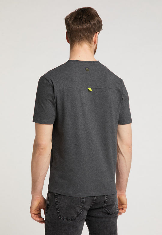 Ein Mann mit kurzen braunen Haaren, der das Recaro_Merch_Daniel T-Shirt Performance in Relaxed-Fit Dunkelgrau mit dezentem RECARO Logo und schwarze Jeans trägt, steht mit dem Rücken zur Kamera vor einem schlichten hellen Hintergrund.