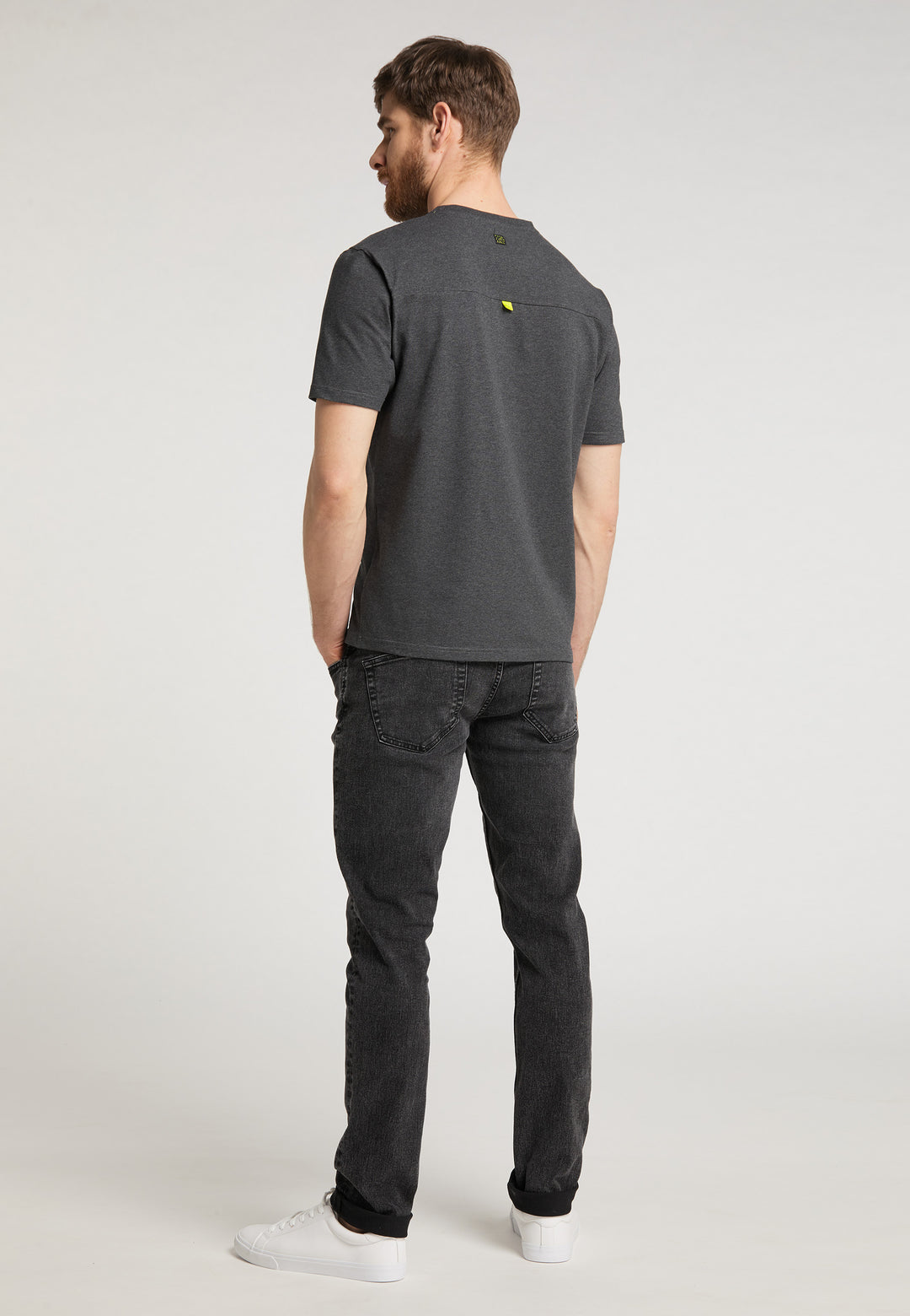 Ein Mann mit kurzen braunen Haaren steht mit abgewandtem Gesicht da und trägt ein locker sitzendes Recaro_Merch_Daniel T-Shirt Performance in Dunkelgrau, schwarze Jeans und weiße Turnschuhe vor einem schlichten hellgrauen Hintergrund.