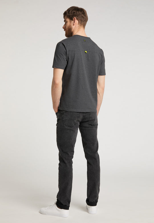 Ein Mann mit kurzen braunen Haaren steht mit abgewandtem Gesicht da und trägt ein locker sitzendes Recaro_Merch_Daniel T-Shirt Performance in Dunkelgrau, schwarze Jeans und weiße Turnschuhe vor einem schlichten hellgrauen Hintergrund.