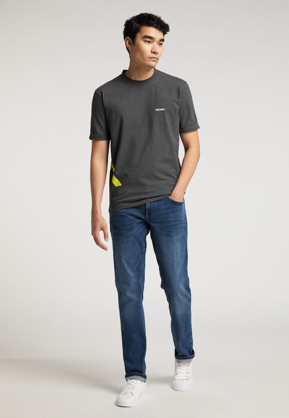 Ein junger Mann mit kurzen dunklen Haaren trägt das RECARO T-Shirt „One Brand One Goal“, blaue Jeans und weiße Turnschuhe. Er steht neben einem RECARO Sportsitz Premium-Gaming-Seat vor einem schlichten hellen Hintergrund, eine Hand in der Tasche.