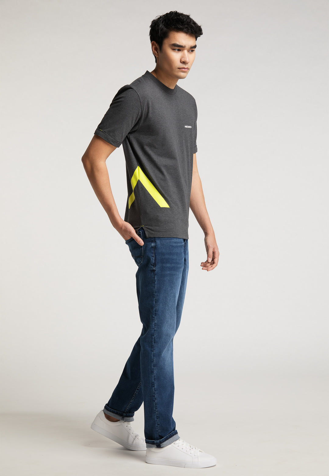 Ein junger Mann steht in einem Studio und trägt das RECARO T-Shirt „One Brand One Goal“ – mit einem gelben geometrischen Design – dazu Bluejeans und weiße Turnschuhe und blickt mit neutralem Gesichtsausdruck zur Seite.