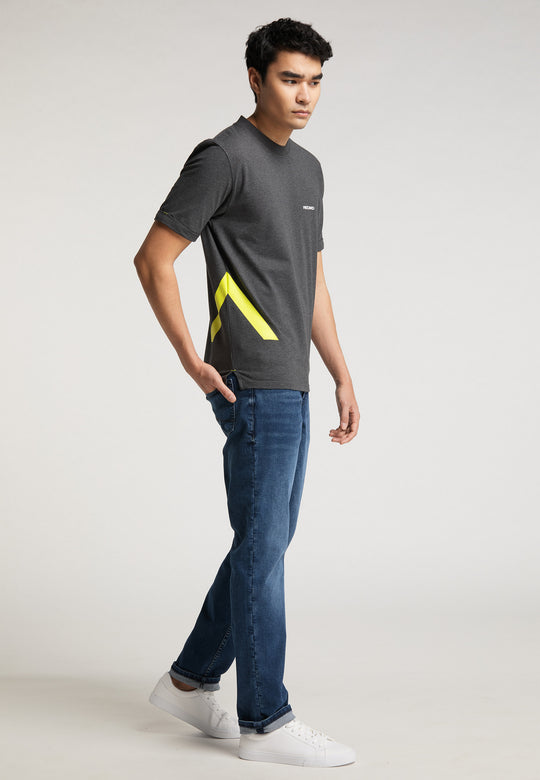 Ein junger Mann steht in einem Studio und trägt das RECARO T-Shirt „One Brand One Goal“ – mit einem gelben geometrischen Design – dazu Bluejeans und weiße Turnschuhe und blickt mit neutralem Gesichtsausdruck zur Seite.