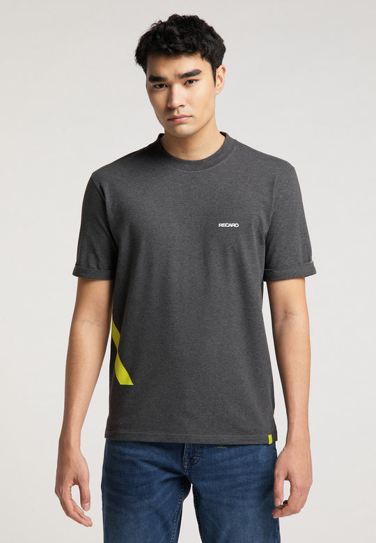 Ein junger Mann mit kurzen schwarzen Haaren und neutralem Gesichtsausdruck blickt nach vorne. Er trägt Bluejeans und das RECARO „T-Shirt One Brand One Goal“ in Dunkelgrau mit gelben Akzenten und einem kleinen Logo, inspiriert vom kultigen Sportsitz-Design.
