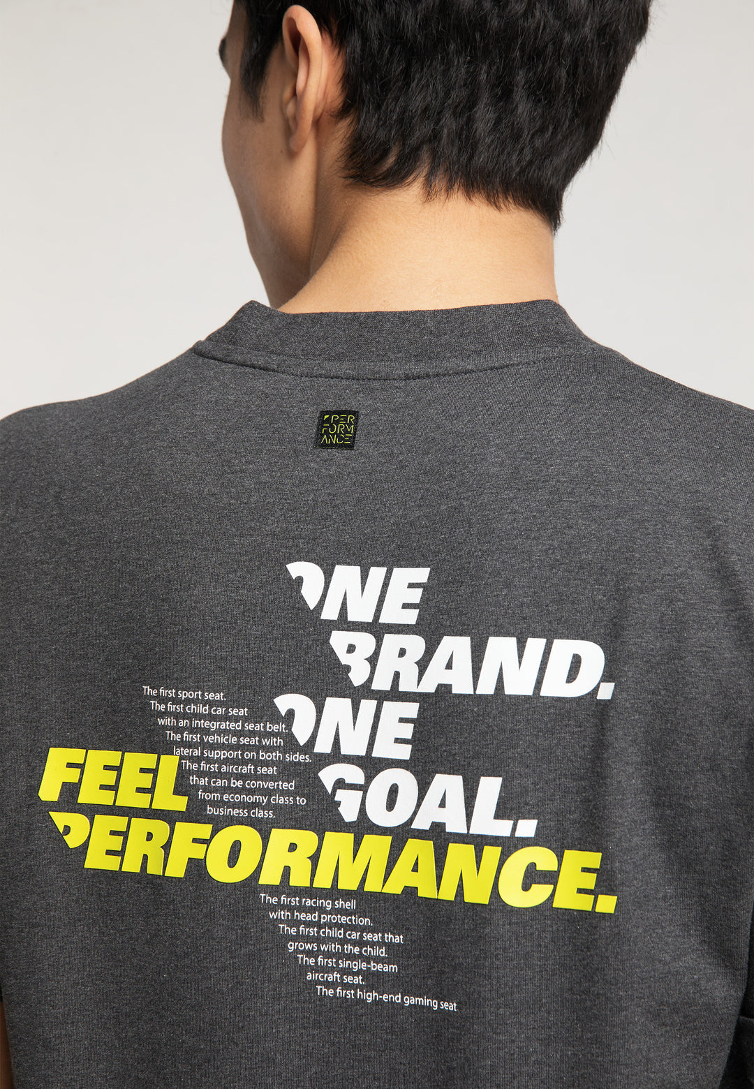 Eine Person trägt das RECARO T-Shirt „One Brand One Goal“ in Dunkelgrau mit dem fettgedruckten weiß-gelben Text „ONE BRAND. ONE GOAL. FEEL PERFORMANCE.“ und einem kleineren weißen „RECARO Premium-Gaming-Seat“-Logo auf der Rückseite.