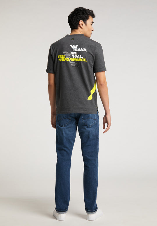 Ein Mann blickt von der Kamera weg und trägt ein RECARO T-Shirt „One Brand One Goal“ – dunkelgrau mit weißem und gelbem Text –, Bluejeans und weiße Turnschuhe vor einem schlichten hellen Hintergrund; ideal für Fans von Premium-Gaming-Seats oder Sportsitzen.