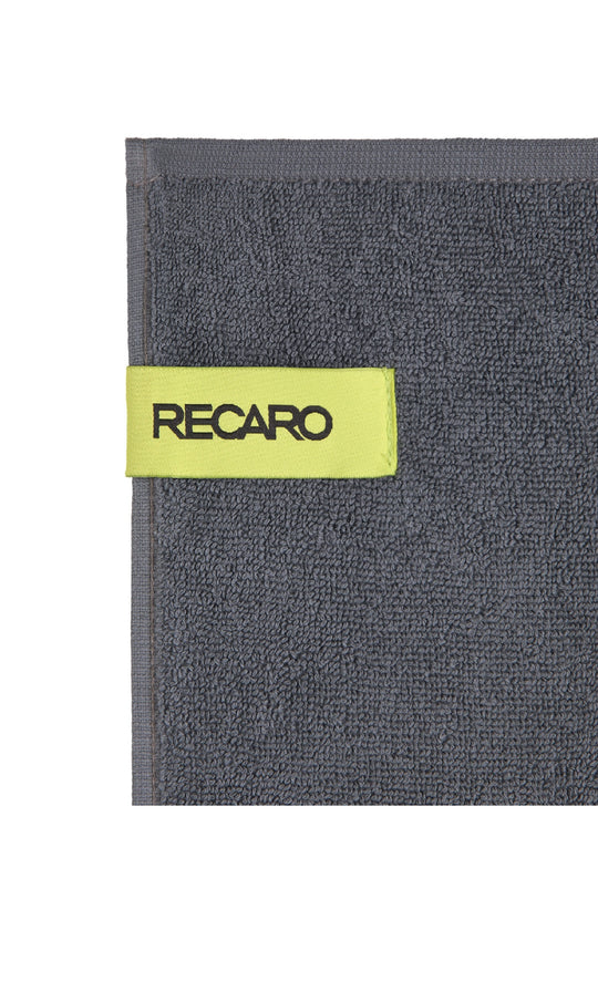 Eine Nahaufnahme eines dunkelgrauen Fitness- und Freizeithandtuchs Performance von RECARO aus hochwertigem Baumwoll-Twill-Frottee mit einer gelbgrünen Stoffschlaufe mit „RECARO“ in schwarzen Buchstaben.