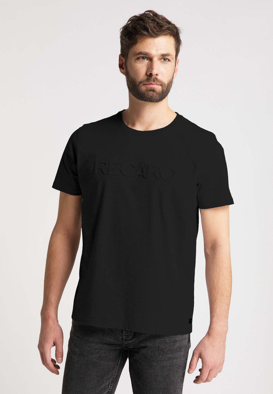 Ein Mann mit kurzen braunen Haaren und Bart trägt ein schwarzes RECARO T-Shirt Originals Embossed aus 100 % Baumwolle mit einem 3D RECARO Logo auf der Vorderseite. Er steht vor einem schlichten hellen Hintergrund und blickt leicht zur Seite.