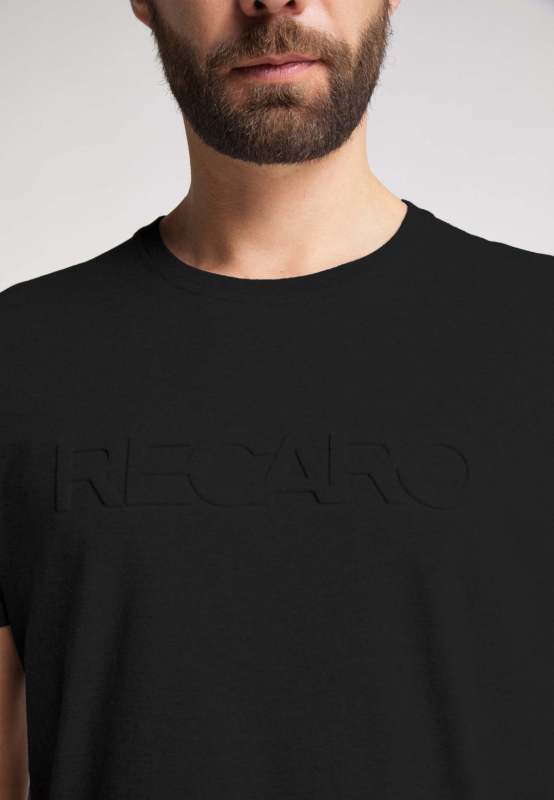 Ein bärtiger Mann trägt das RECARO T-Shirt Originals Embossed aus 100 % Baumwolle mit einem auffälligen 3D RECARO Logo auf der Brust. Der Hintergrund ist schlicht und neutral.