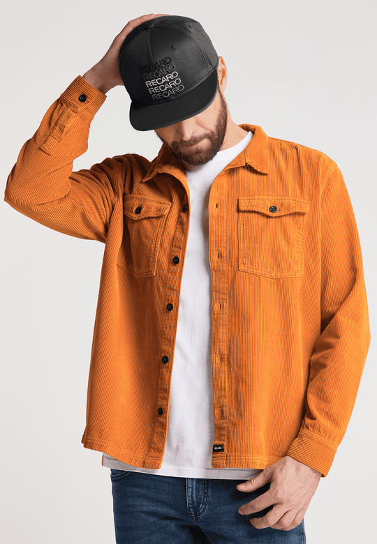 Ein Mann, der die RECARO_Merchandising Snapback Cap Spektrum trägt, ein orangefarbenes Cordhemd über einem weißen T-Shirt und blaue Jeans, steht vor einem schlichten Hintergrund, neigt den Kopf nach unten und berührt den Schirm der Kappe.