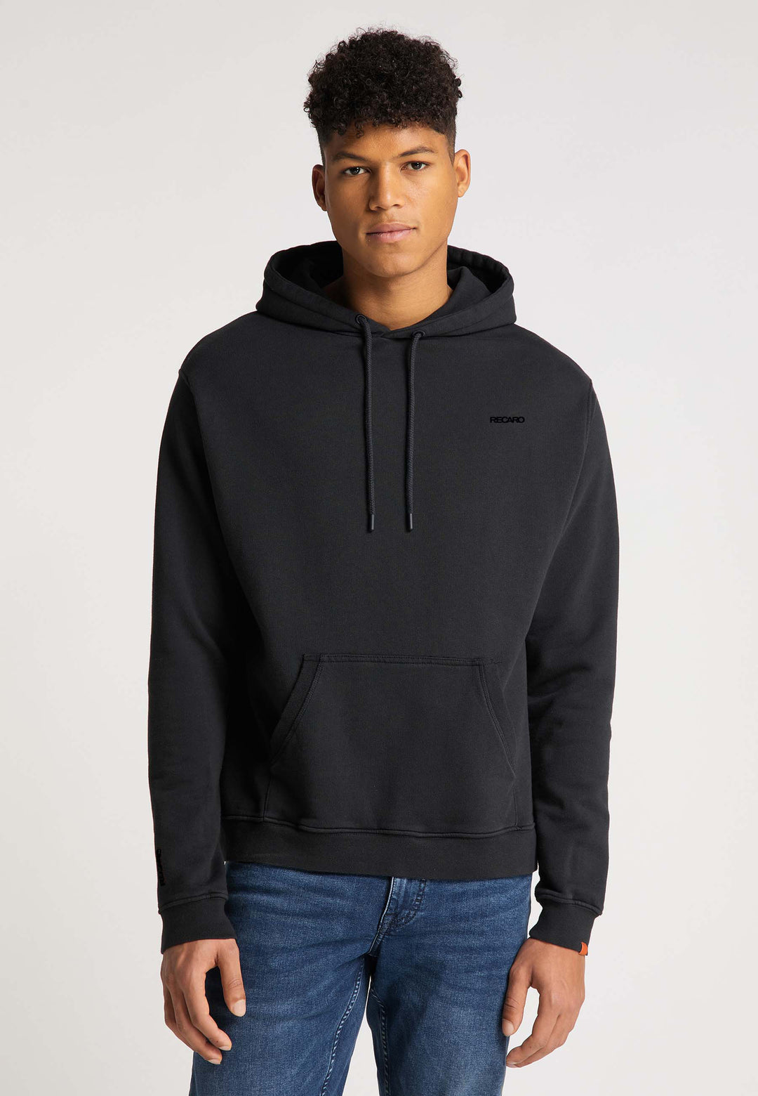 Ein junger Mann mit kurzen lockigen Haaren trägt den schwarzen Recaro Hoodie Originals Basic und Blue Jeans und steht vor einem schlichten hellen Hintergrund.