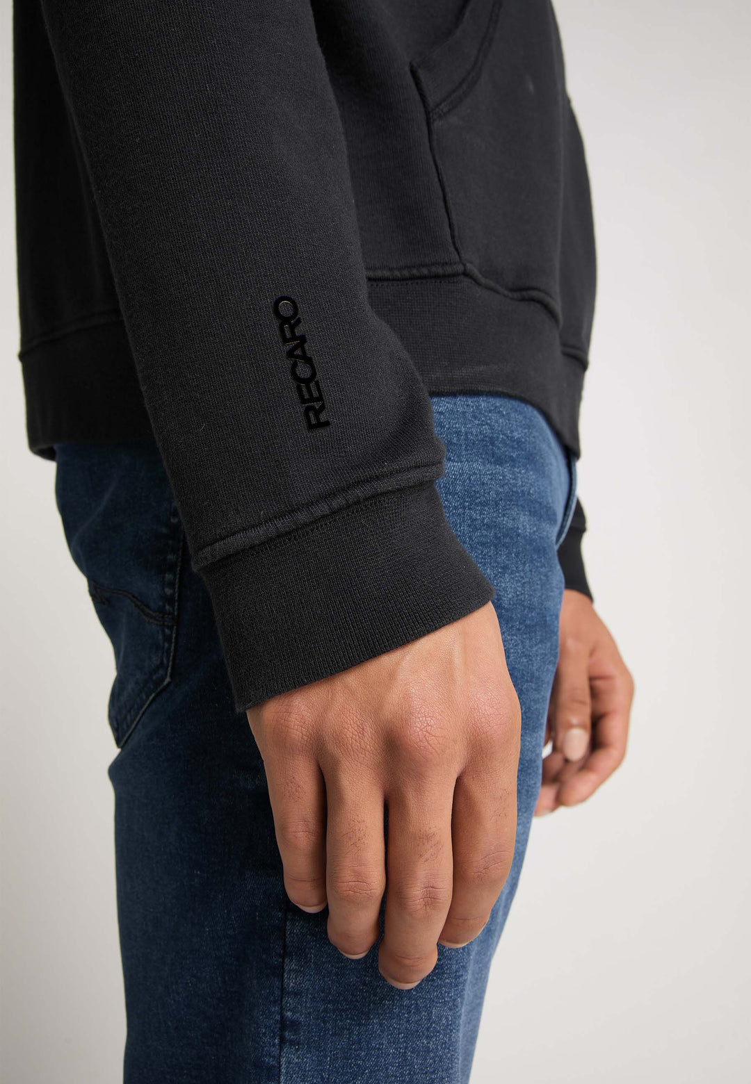 Eine Person trägt den schwarzen Recaro Hoodie Originals Basic aus weicher Baumwolle mit RECARO-Stickerei auf dem Ärmel, kombiniert mit Bluejeans; von der Taille abwärts gezeigt, mit sichtbarem linken Arm und Hand.