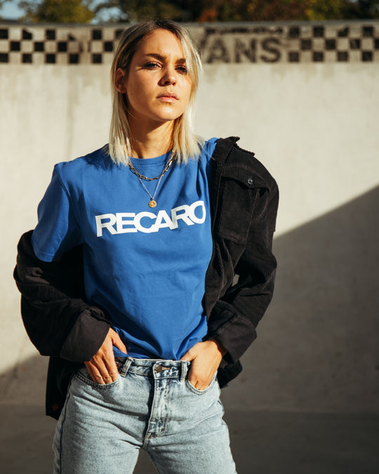 Eine blonde Frau steht selbstbewusst im Freien in einem blauen RECARO T-Shirt Originals, mit einer schwarzen Jacke über den Schultern und hellblauen Jeans. Hinter ihr ist eine sonnenbeschienene Betonwand mit Schachbrettmuster.