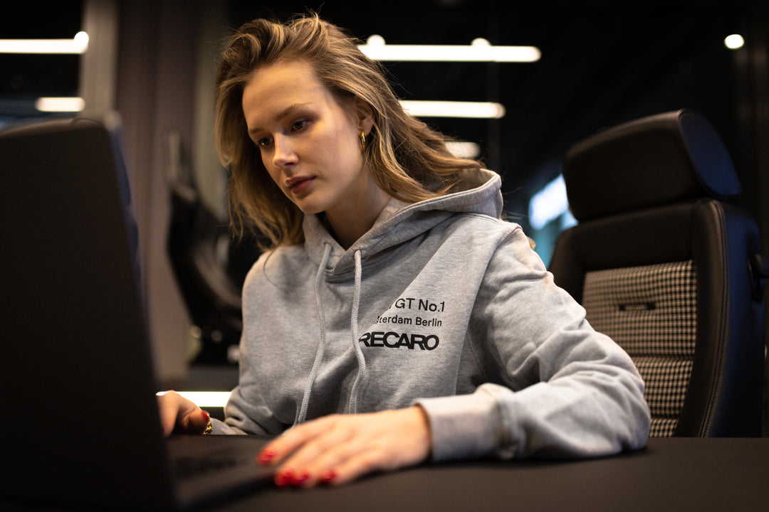 Eine Frau trägt den LMG x RECARO Hoodie von Recaro, während sie in einem modernen Büro an ihrem Laptop arbeitet und so dezent den Stil des LMG Racing Club und der GT4 European Series zur Schau stellt. Vor dem dunklen Hintergrund sind Bürostühle zu sehen.
