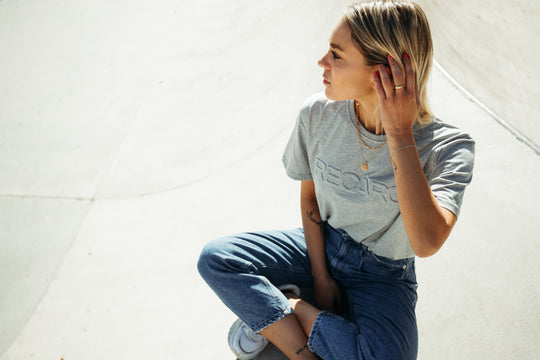 Eine Person mit blondem Haar sitzt im Schneidersitz auf einer sonnenbeschienenen Fläche. Sie trägt ein graues RECARO  Originals Embossed, Blue Jeans und weiße Turnschuhe und blickt nachdenklich zur Seite, während sie sich durch die Haare fährt.