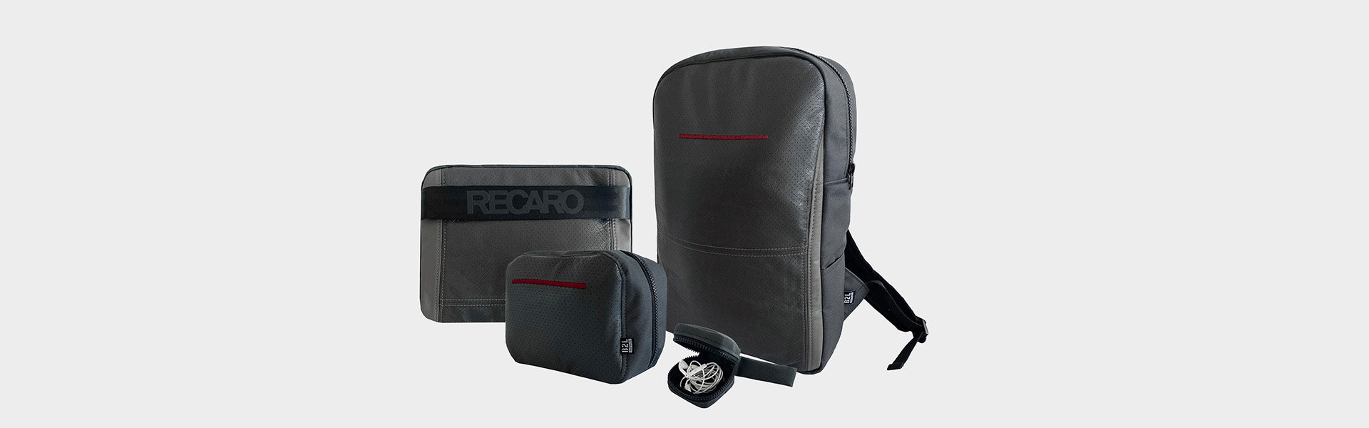 Drei graue Reisetaschen unterschiedlicher Größe, darunter ein Rucksack, eine mittelgroße Tasche mit schwarzem RECARO-Gurt und eine kleine Tasche. Vorne befindet sich ein schwarzer Gürtel mit silberner Rundschnalle. Alle Artikel haben minimale rote Akzente.