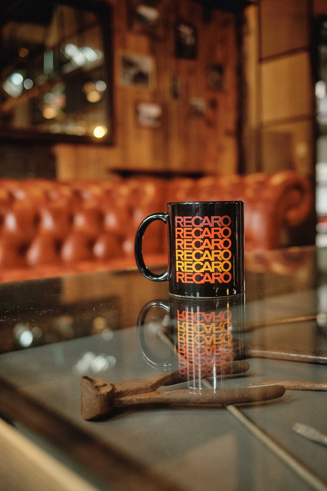 Eine RECARO_Merchandising Tasse Spektrum mit rot-gelber Aufschrift steht auf einem Glastisch neben einer Zange, während ein braunes Ledersofa und eine holzgetäfelte Wand mit gerahmten Fotos die Atmosphäre des Spektrum Designs verstärken.