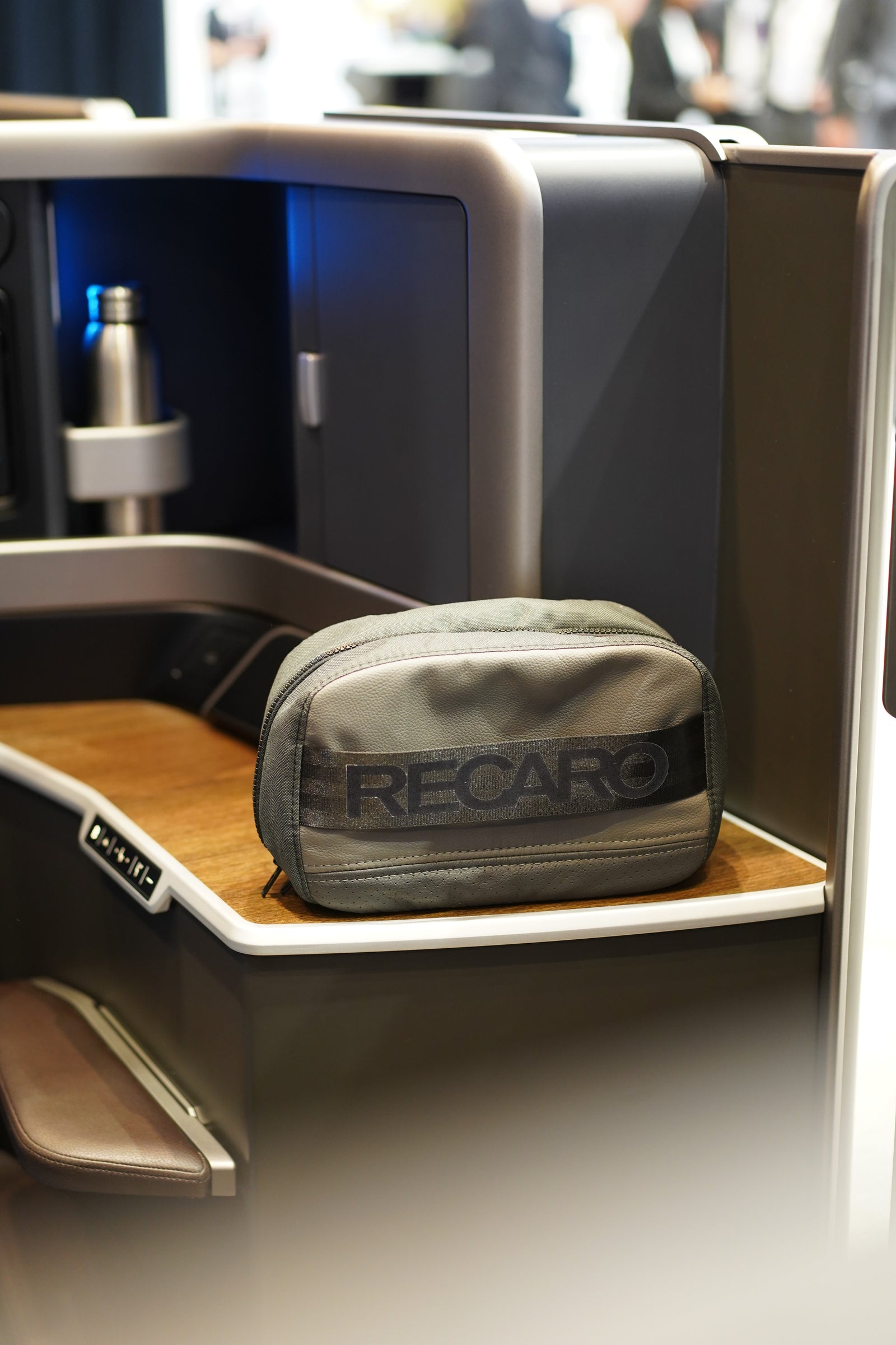 Eine graue Reisetasche mit RECARO-Logo liegt auf der hölzernen Sitzkonsole eines Business-Class-Flugzeugs, neben einer Wasserflasche aus Metall und einer gebogenen Trennwand.