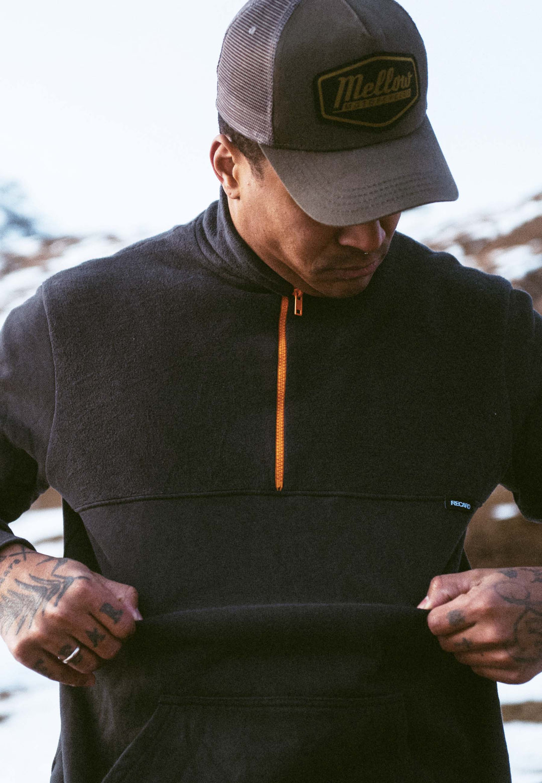 Eine Person in Freizeitkleidung, die das Recaro_Merch_Daniel Half Zip Sweatshirt Originals und eine Baseballkappe trägt, blickt nach unten, während sie den unteren Teil ihres Fleece festhält; Schnee und Berge verschwimmen im Hintergrund.