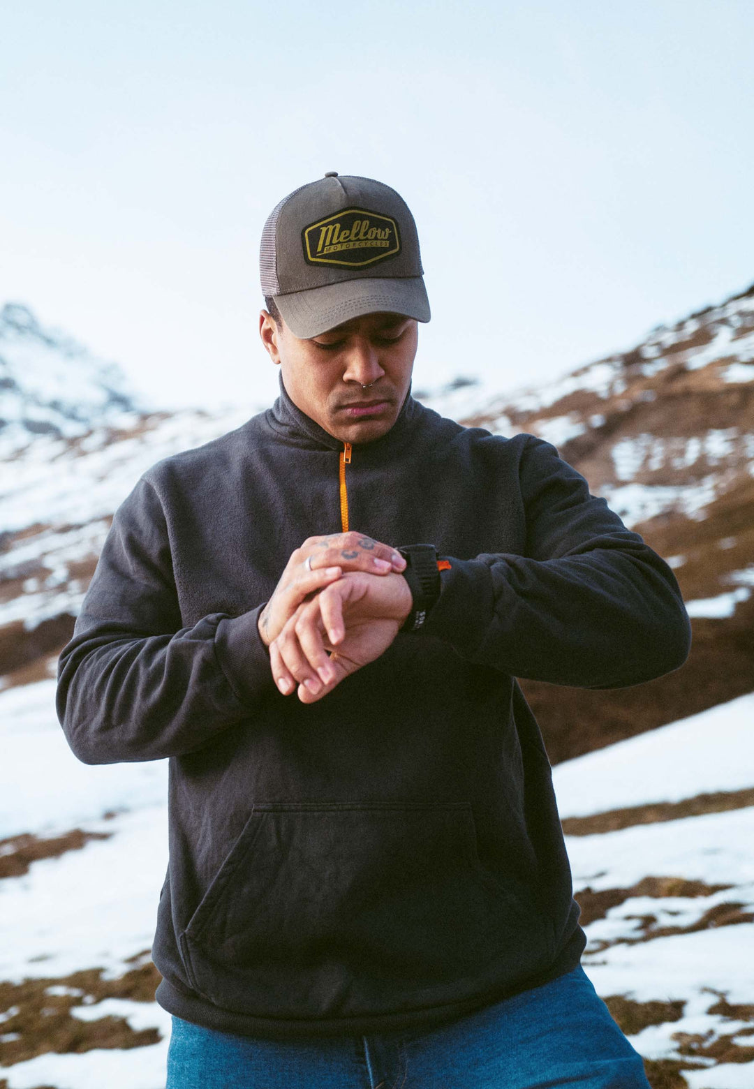 Ein Mann mit dunklem Kapuzenpullover und grauer Mütze steht draußen in den schneebedeckten Bergen. Er trägt das Half Zip Sweatshirt Originals von Recaro_Merch_Daniel und schaut nach unten, um auf die Uhr zu schauen.