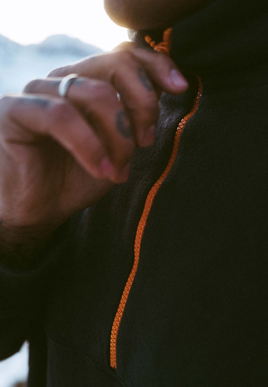 Eine Nahaufnahme zeigt eine Hand, die den orangefarbenen Reißverschluss des RECARO Half Zip Sweatshirt Originals schließt. Ein Ring ist sichtbar, und verschwommene Berge und Himmel deuten auf Outdoor-Training oder Freizeitkleidung hin.