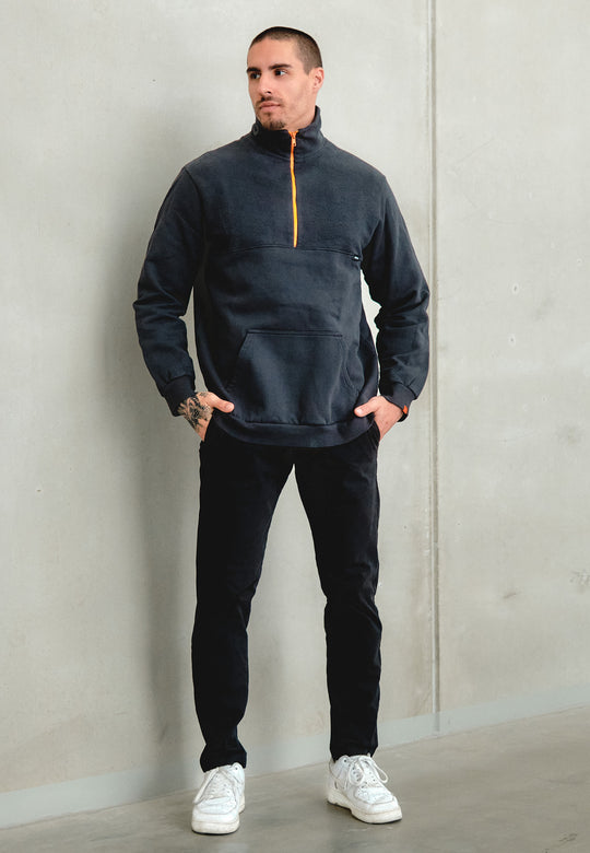 Ein Mann mit kurzen Haaren, Bart und einer Tätowierung auf der linken Hand steht mit den Händen in den Taschen vor einer hellgrauen Wand und trägt das Recaro_Merch_Daniel Half Zip Sweatshirt Originals (dunkel mit orangefarbenem Reißverschluss), eine schwarze Hose und weiße Turnschuhe.