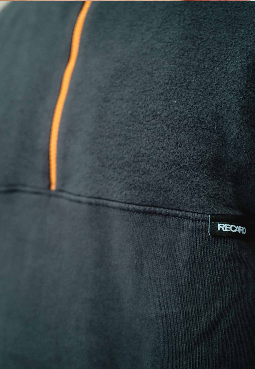 Nahaufnahme eines schwarzen Half Zip Sweatshirt Originals von Recaro_Merch_Daniel mit orangefarbener Naht und kleinem schwarzen RECARO-Etikett. Der strukturierte Stoff eignet sich sowohl für Outdoor-Training als auch für die Freizeit.