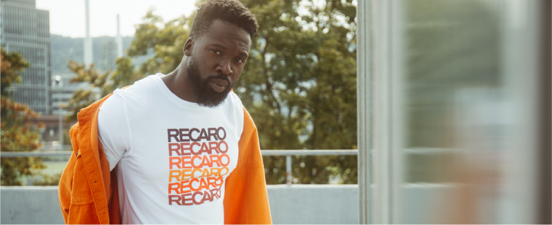 Ein Mann mit Bart trägt ein weißes T-Shirt mit dem Schriftzug RECARO in Rot und Schwarz und eine orangefarbene Jacke über den Schultern. Er steht im Freien, im Hintergrund sind Bäume und Gebäude zu sehen.