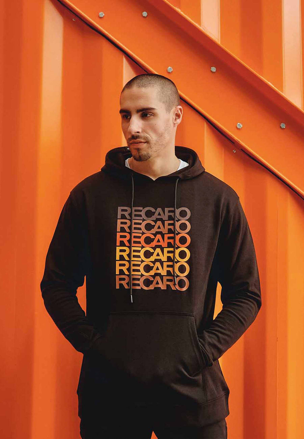 Ein Mann mit kurzen Haaren und gestutztem Bart trägt den schwarzen RECARO Hoodie Spektrum mit orangefarbenem und gelbem Text und steht vor einer orangefarbenen Metallwand mit diagonalen Balken für einen coolen Retro-Look.