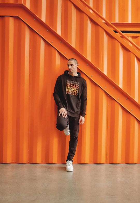 Eine Person trägt den RECARO Hoodie Spektrum und weiße Turnschuhe und steht mit einem erhobenen Fuß vor einer orangefarbenen Wellblechwand und Treppe und blickt zur Seite.